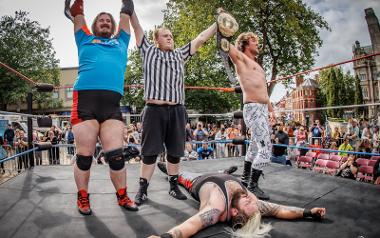 Two wrestlers hold up trophy belts after a match in a wrestling ring on the Flag Market with crowds cheering