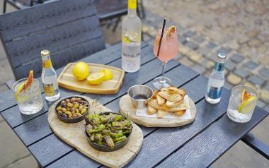 A table of cocktails and wooden slabs with plates of roasted olives, peppers and bread outside Forum Bar and Kitchen