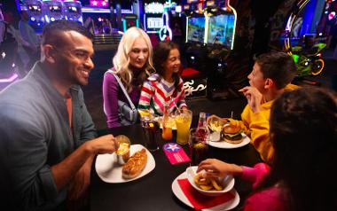 A family sat at a table enjoying hot dogs and burgers with arcade machines in the background