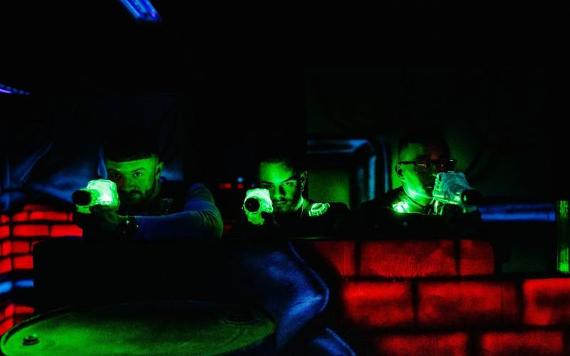 Three people pointing laser tag guns through a gap in wall of arena.