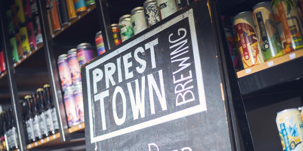 Priest Town Brewing blackboard sign, with beers on shelves behind inside shop