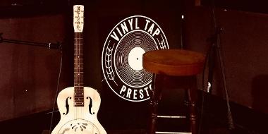 Guitar, bar stool, and Vinyl Tap Preston logo ready for music performance.