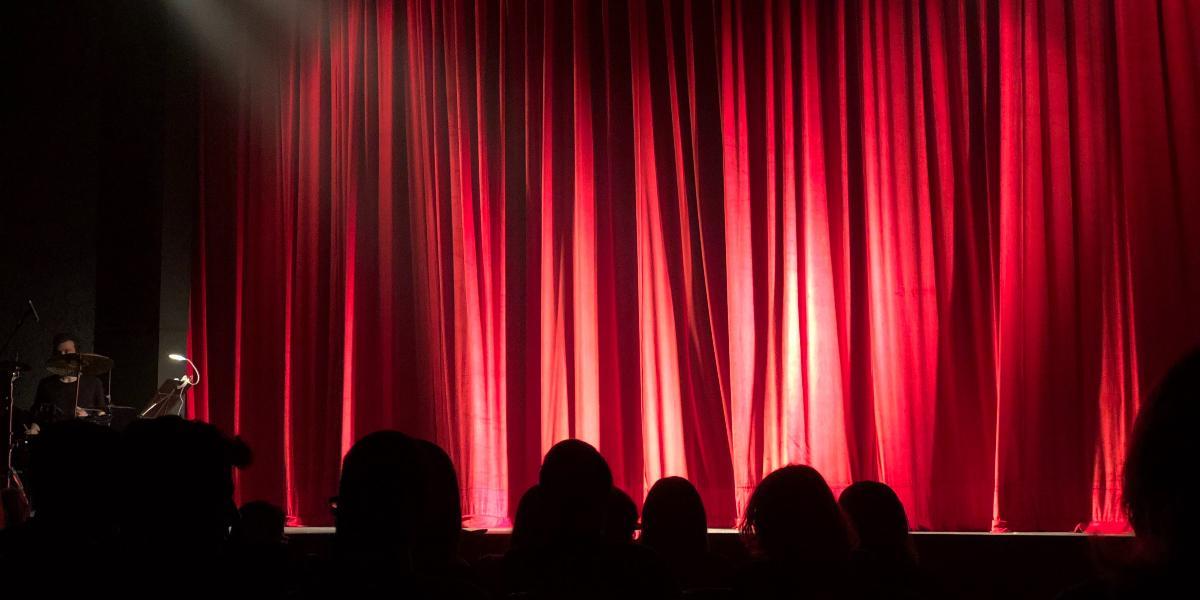 Audience sat in theatre with red curtain closed on stage.