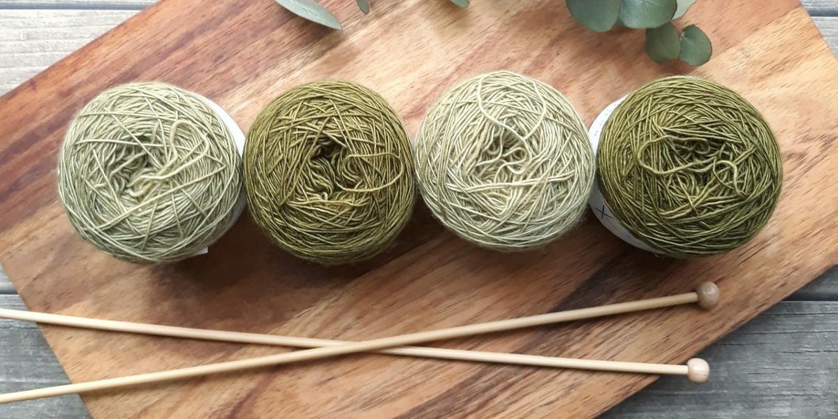 Different shades of balls of wool arranged in row on wooden board with knitting needles beside them.