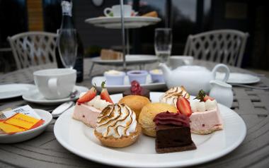 Array of cakes on a plate with afternoon tea stand and bottle of Prosecco in the background.